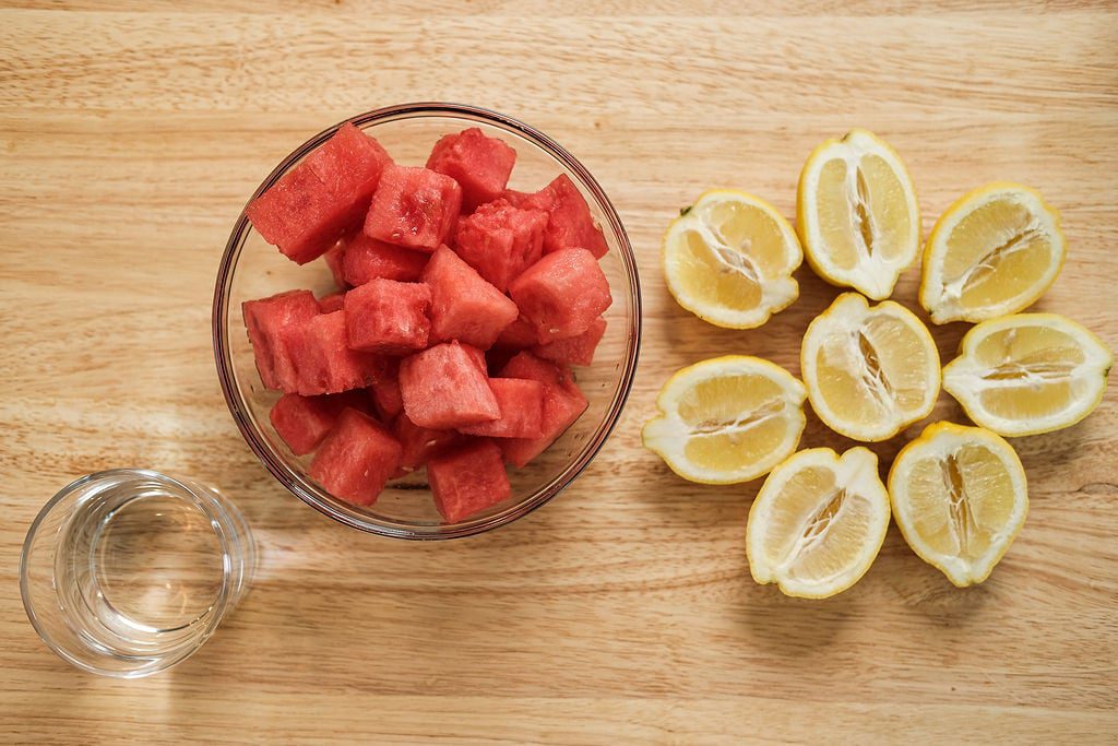 Clear class of water next to clear glass bowl of sliced watermelon next to sliced lemons on top of light wood table.