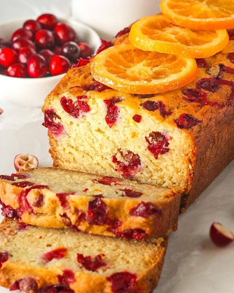 Loaf of cranberry bread with 2 slices cut off and laying down with orange slices on top next to bowl of fresh cranberries.